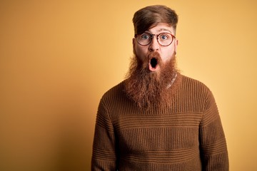 Canvas Print - Handsome Irish redhead man with beard wearing glasses and winter sweater over yellow background afraid and shocked with surprise expression, fear and excited face.