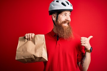 Wall Mural - Redhead Irish delivery man with beard wearing bike helmet and holding takeaway paper bag pointing and showing with thumb up to the side with happy face smiling