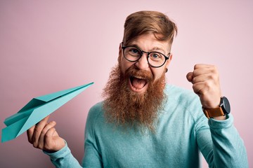 Poster - Irish redhead man with beard holding paper plane over pink isolated background screaming proud and celebrating victory and success very excited, cheering emotion