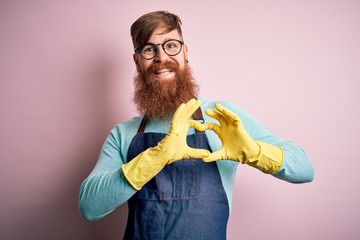 Canvas Print - Irish redhead housekeeping man with beard wearing apron and washing gloves smiling in love showing heart symbol and shape with hands. Romantic concept.