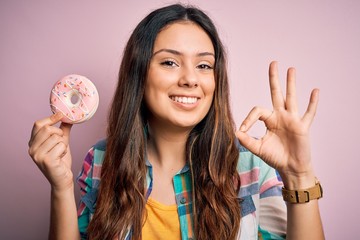 Young beautiful brunette woman eating sweet pink doughnut over isolated background doing ok sign with fingers, excellent symbol