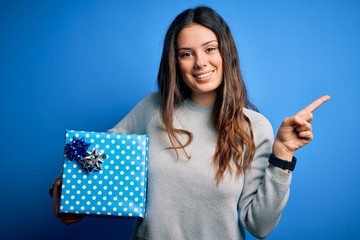 Wall Mural - Young beautiful brunette woman holding birthday gift over isolated blue background very happy pointing with hand and finger to the side