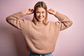 Poster - Young beautiful blonde woman wearing winter wool sweater over pink isolated background Smiling pulling ears with fingers, funny gesture. Audition problem