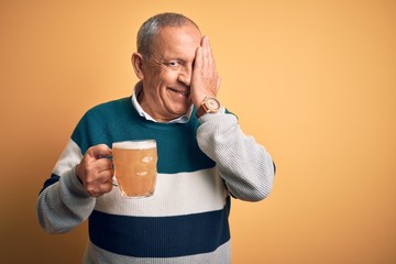 Sticker - Senior handsome man drinking jar of beer standing over isolated yellow background covering one eye with hand, confident smile on face and surprise emotion.