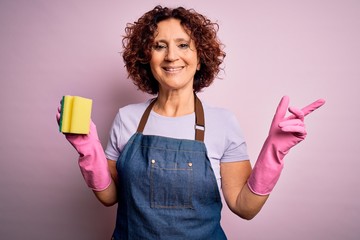 Poster - Middle age curly hair woman cleaning doing housework wearing apron and gloves using scourer very happy pointing with hand and finger to the side