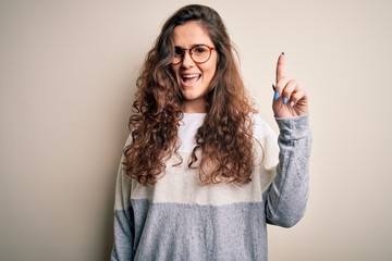 Sticker - Young beautiful woman with curly hair wearing sweater and glasses over white background pointing finger up with successful idea. Exited and happy. Number one.