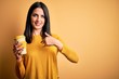 © Krakenimages.com - Young woman with blue eyes holding cup of coffee standing over yellow background with surprise face pointing finger to himself