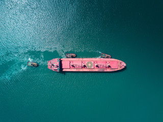 Aerial drone shoot the cargo ship enters the international trade sea port. The cargo ship sails accompanied by three port tugs. Top view