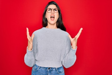 Canvas Print - Young beautiful brunette woman wearing casual sweater over red background crazy and mad shouting and yelling with aggressive expression and arms raised. Frustration concept.