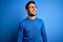 Young Handsome Man With Beard Wearing Casual Sweater And Glasses Over Blue Background Looking Away To Side With Smile On Face, Natural Expression. Laughing Confident.