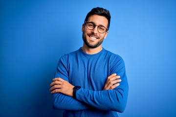 Wall Mural - Young handsome man with beard wearing casual sweater and glasses over blue background happy face smiling with crossed arms looking at the camera. Positive person.