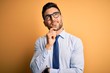 © Krakenimages.com - Young handsome businessman wearing tie and glasses standing over yellow background looking confident at the camera smiling with crossed arms and hand raised on chin. Thinking positive.