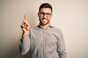 Young handsome man wearing elegant shirt and glasses over isolated white background showing and pointing up with finger number one while smiling confident and happy.