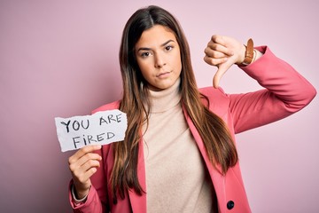 Poster - Young beautiful girl holding paper with you are fired message over isolated pink background with angry face, negative sign showing dislike with thumbs down, rejection concept