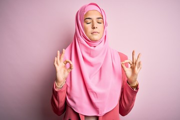 Poster - Young beautiful girl wearing muslim hijab standing over isolated pink background relaxed and smiling with eyes closed doing meditation gesture with fingers. Yoga concept.