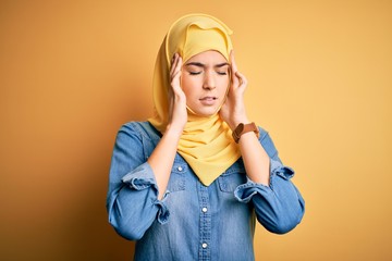 Wall Mural - Young beautiful girl wearing muslim hijab standing over isolated yellow background with hand on head for pain in head because stress. Suffering migraine.