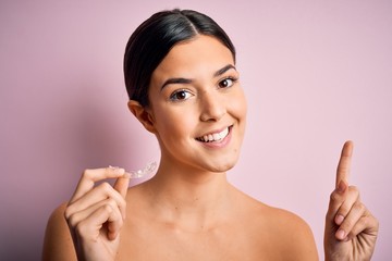 Poster - Young beautiful girl holding dental aligner over isolated pink background surprised with an idea or question pointing finger with happy face, number one