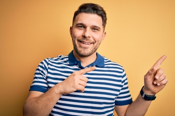 Young man with blue eyes wearing casual striped t-shirt over yellow background smiling and looking at the camera pointing with two hands and fingers to the side.