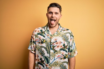 Wall Mural - Young man with blue eyes on vacation wearing floral summer shirt over yellow background sticking tongue out happy with funny expression. Emotion concept.