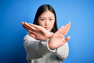 Canvas Print - Young beautiful asian woman wearing casual sweater standing over blue isolated background Rejection expression crossing arms and palms doing negative sign, angry face