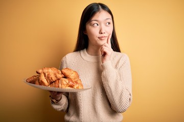 Canvas Print - Young asian woman holding french pastry croissant over yellow isolated background serious face thinking about question, very confused idea