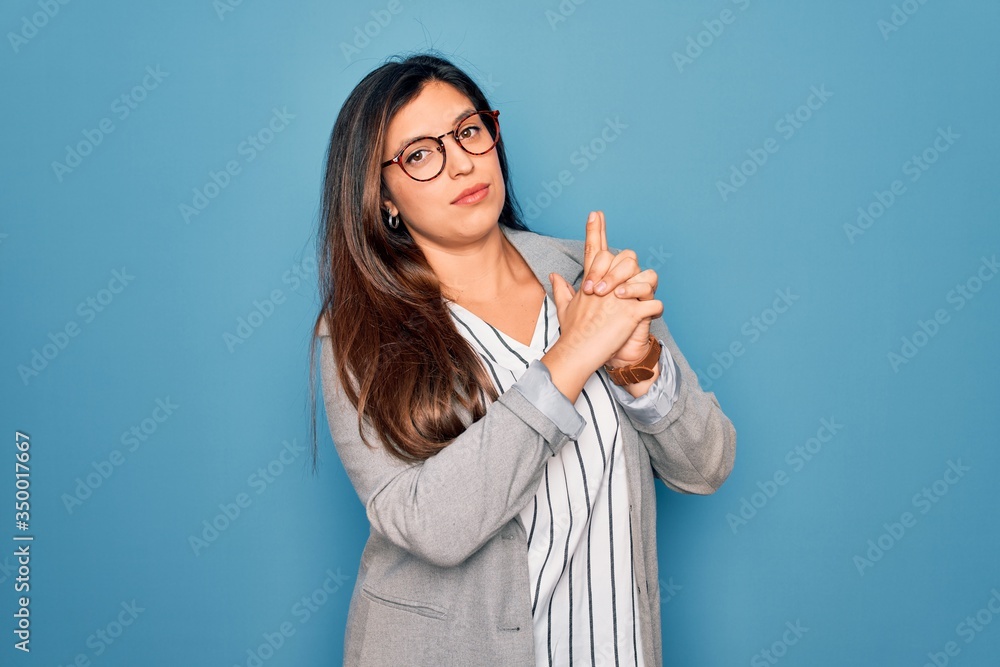 Young hispanic business woman wearing glasses standing over blue isolated background Holding symbolic gun with hand gesture, playing killing shooting weapons, angry face