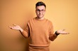 © Krakenimages.com - Young handsome caucasian man wearing glasses and casual winter sweater over yellow background clueless and confused expression with arms and hands raised. Doubt concept.