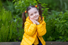 Cute Kid Girl With Ponytails In A Yellow Jacket Sits On A Bench And Smiles