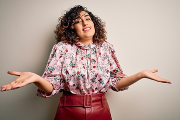 Poster - Young beautiful curly arab woman wearing floral t-shirt standing over isolated white background clueless and confused expression with arms and hands raised. Doubt concept.