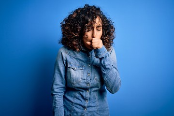 Poster - Young beautiful curly arab woman wearing casual denim shirt standing over blue background feeling unwell and coughing as symptom for cold or bronchitis. Health care concept.