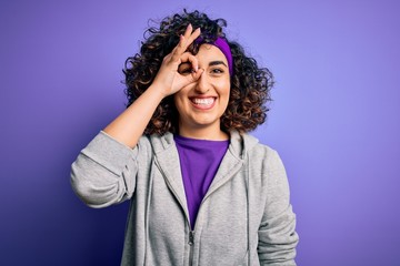 Sticker - Beautiful curly arab sportswoman doing sport wearing sportswear over purple background doing ok gesture with hand smiling, eye looking through fingers with happy face.