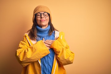 Sticker - Middle age woman wearing yellow raincoat and winter hat over isolated background smiling with hands on chest with closed eyes and grateful gesture on face. Health concept.