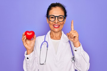 Sticker - Middle age senior cardiologist doctor woman holding red heart over purple background surprised with an idea or question pointing finger with happy face, number one