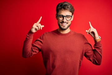 Wall Mural - Young handsome man with beard wearing glasses and sweater standing over red background smiling pointing to head with both hands finger, great idea or thought, good memory