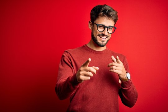 young handsome man with beard wearing glasses and sweater standing over red background pointing fing