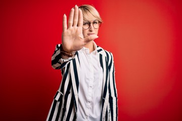 Poster - Blonde business woman with short hair wearing glasses and striped jacket over red background doing stop sing with palm of the hand. Warning expression with negative and serious gesture on the face.