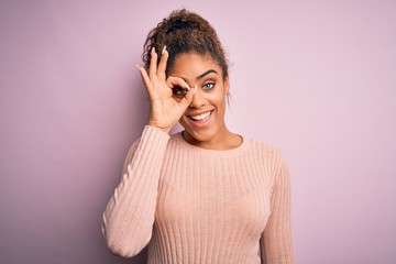 Canvas Print - Young beautiful african american girl wearing casual sweater standing over pink background doing ok gesture with hand smiling, eye looking through fingers with happy face.