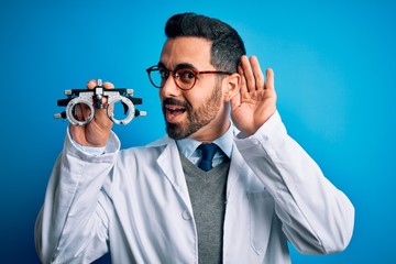 Sticker - Young handsome optical man with beard holding optometry glasses over blue background smiling with hand over ear listening an hearing to rumor or gossip. Deafness concept.