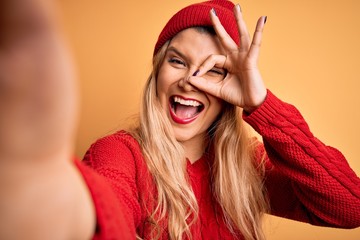 Poster - Young beautiful blonde woman making selfie by camera over isolated yellow background with happy face smiling doing ok sign with hand on eye looking through fingers