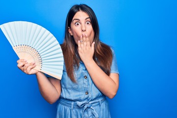 Sticker - Young beautiful brunette woman holding wooden hand fan over isolated blue background covering mouth with hand, shocked and afraid for mistake. Surprised expression