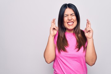 Sticker - Young beautiful brunette woman wearing casual pink t-shirt standing over white background gesturing finger crossed smiling with hope and eyes closed. Luck and superstitious concept.