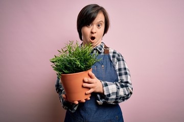 Wall Mural - Young down syndrome gardener woman wearing worker apron holding green plant pot scared in shock with a surprise face, afraid and excited with fear expression