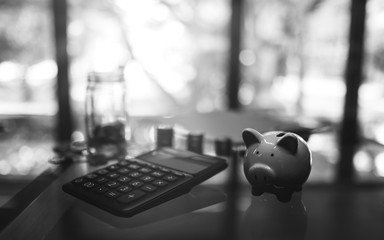 Black and white image of a calculator , piggy bank , coins stack and a glass money jar on the table for saving and financial concept