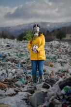 Woman With Gas Mask Holding Green Plant On Landfill, Environmental Concept.