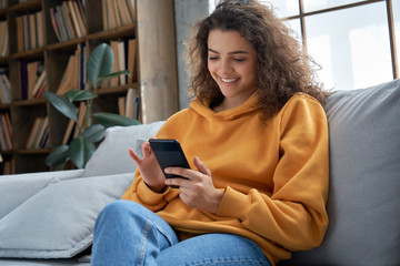 happy millennial hispanic teen girl checking social media holding smartphone at home. smiling young 