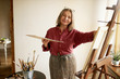 © shurkin_son - Creativity, inspiration, art and painting concept. Portrait of positive talented middle aged female artist wearing apron standing in front of easel, holding brush and palette, working in her studio