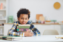 Portrait Of Cute African-American Boy Holding Stack Of Books While Sitting At Desk At Home And Smiling At Camera, Copy Space