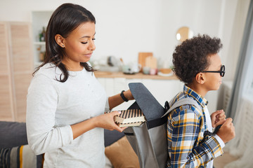 Wall Mural - Side view portrait of smiling African-American woman putting books into backpack while sending son to school