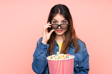 Young brunette woman over isolated pink background surprised with 3d glasses and holding a big bucket of popcorns