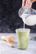 A milk is pouring into a glass with premium organic japan matcha tea on gray table for modern lifestyle latte preparation. Adding a hot milk foam milk on top in green tea